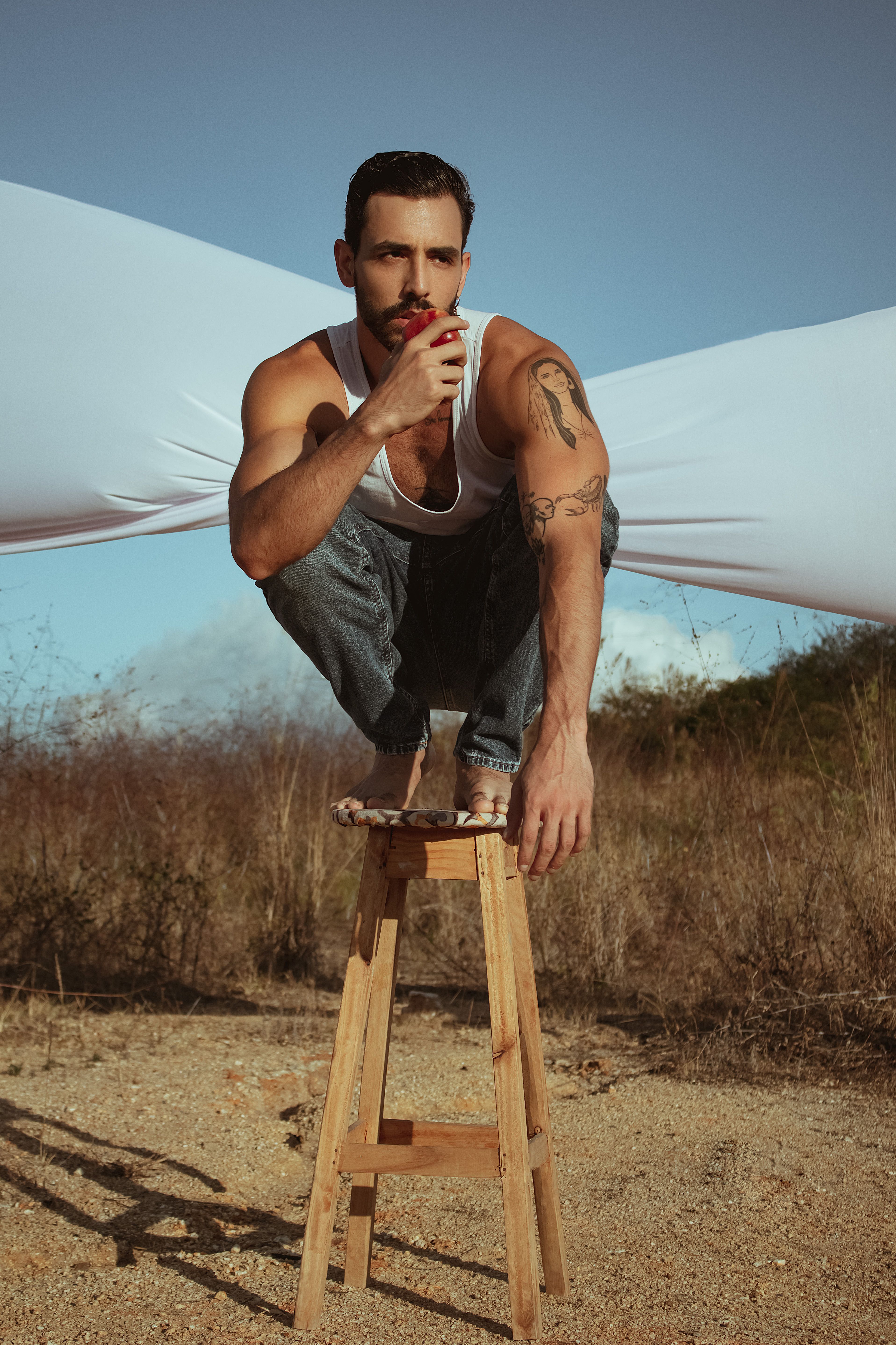 A fashionable man crouches on a stool outdoors, wearing jeans and a tank top.