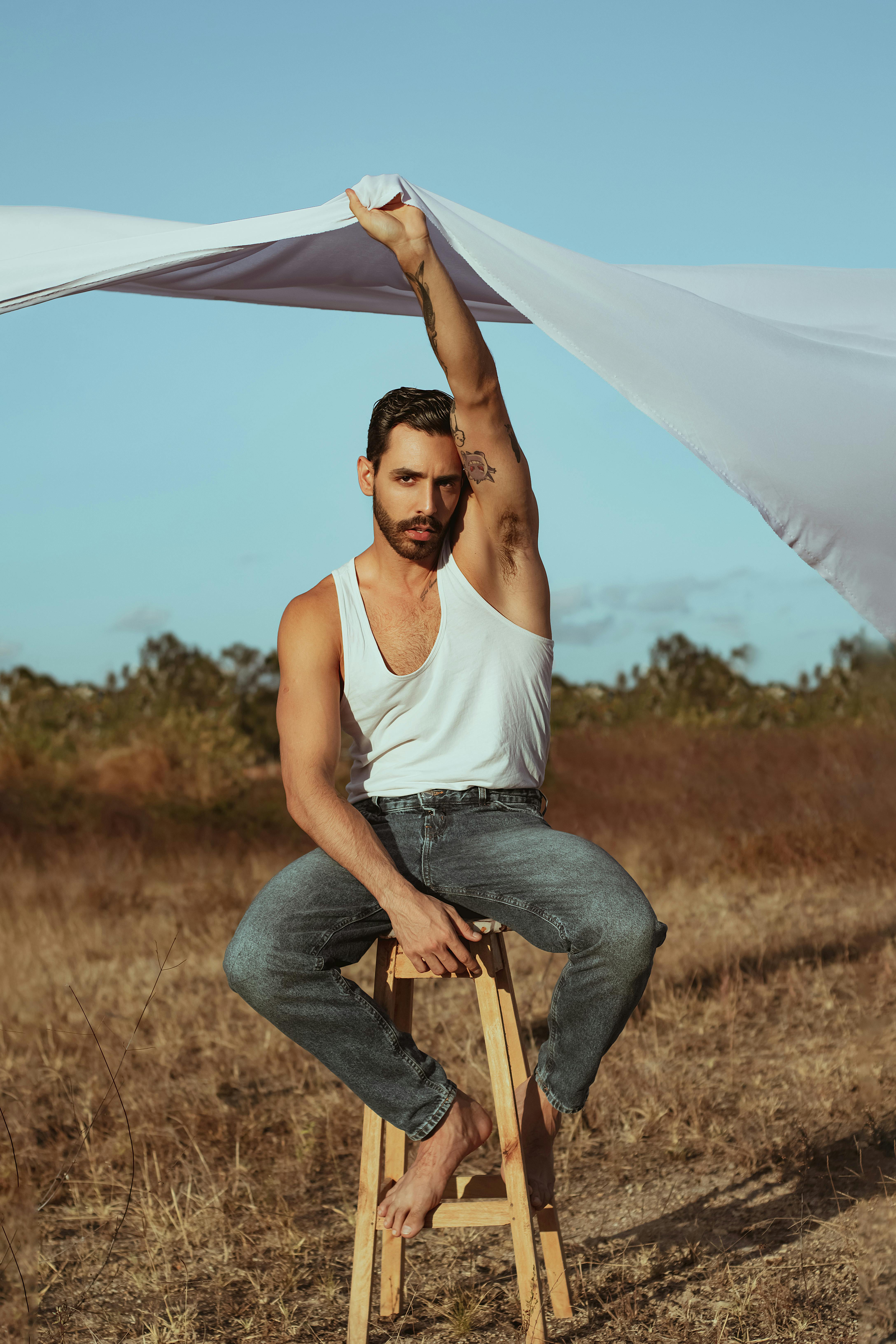 Man in jeans on a stool outdoors with fabric overhead, showcasing fashion and style.