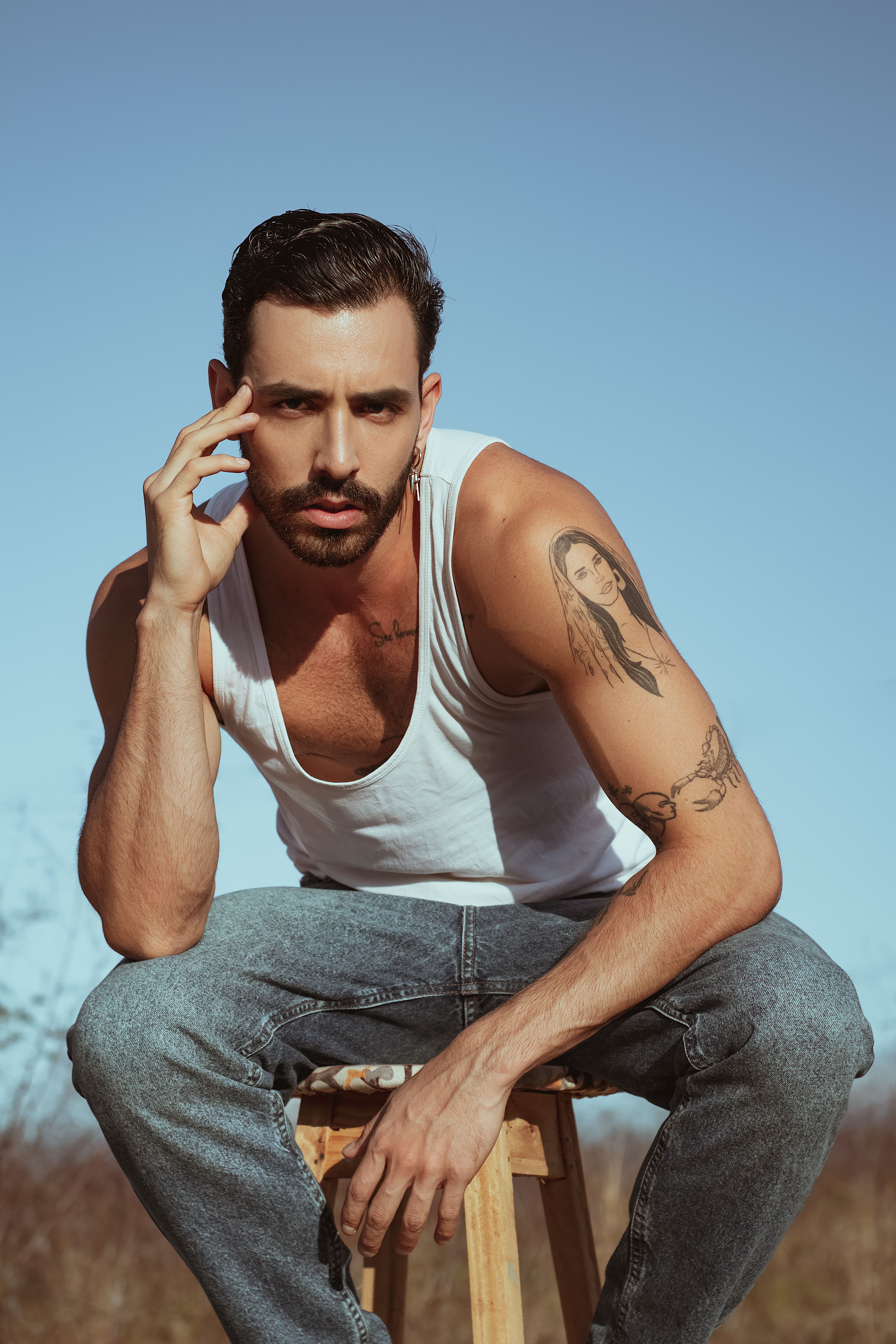 Young man in a tank top posing outdoors with a thoughtful expression and visible tattoo.