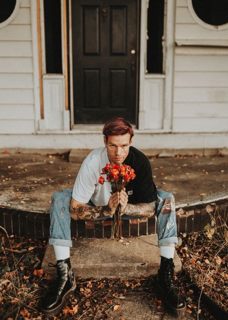 Tattooed man in casual attire holding red roses, sitting in front of an abandoned house in Des Moines.