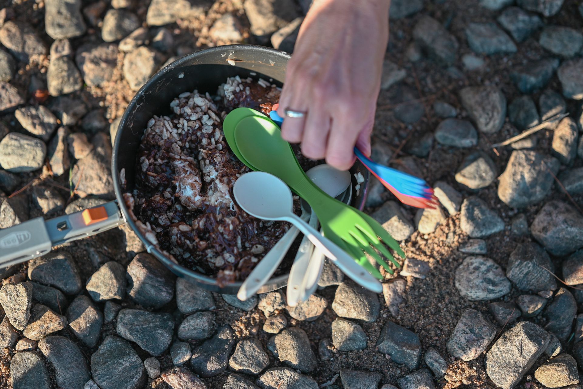 A close-up of a camping cooking pot with rice and utensils on a rocky surface outdoors.