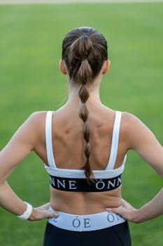 Back view of a woman with hands on hips and braided hair, wearing a sports bra, standing outdoors.
