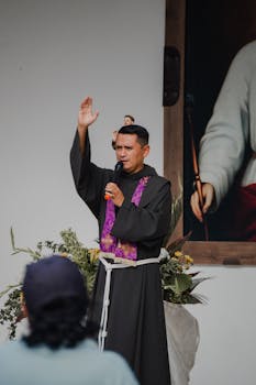 A priest in clerical clothing raises his hand in blessing during an outdoor religious ceremony.