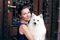 在户外，一位穿着蓝色连衣裙、面带微笑的女子拥抱着一只白色的萨摩耶狗