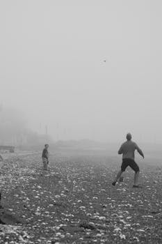 A father and child enjoy playtime on a foggy beach in black and white.