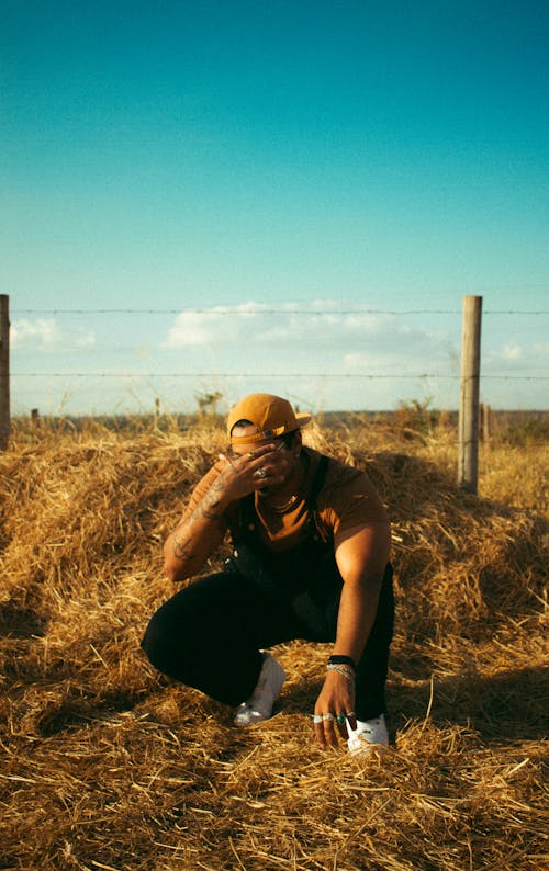 Man in Black T-shirt and Brown Cap Sitting on Brown Grass Field Covering His Face