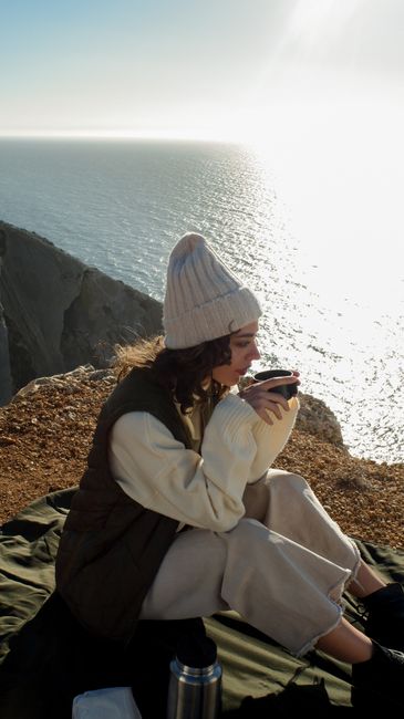 Woman sitting on a cliff with a drink, enjoying the ocean view during sunset.