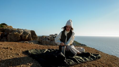 Free Young woman with thermos enjoys picnic by the ocean on a sunny day. Stock Photo