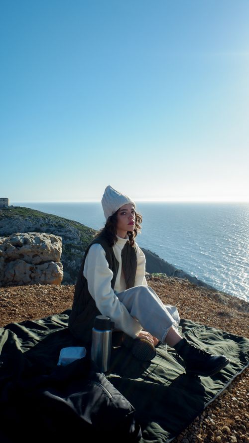 Free A woman sits by the ocean enjoying a peaceful sunset, wrapped in warm clothing. Stock Photo