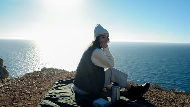 A woman relaxes on a clifftop, overlooking the ocean during sunset, capturing a serene moment.