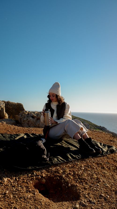 Free Woman with a knit hat sits outdoors enjoying a hot beverage with a scenic mountain view. Stock Photo