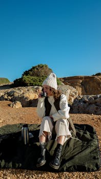 Woman in beanie drinking outdoors on a sunny day, sitting on blanket among rocks.