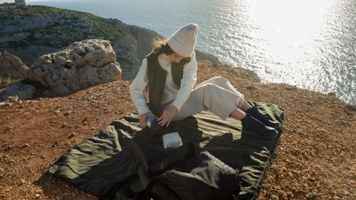 Free A woman in a knit hat enjoying a peaceful picnic on a cliffside with a scenic ocean view. Stock Photo