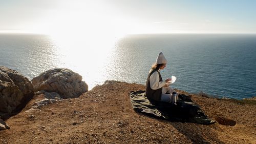 Free A woman in a beanie sits on a cliffside in Lisbon, enjoying a sunny ocean view. Stock Photo