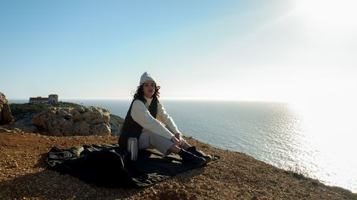 Free A woman in winter clothing sits on a cliff in Lisbon, overlooking the ocean under a bright sky. Stock Photo