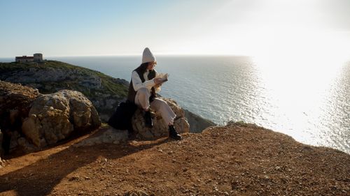Free Woman enjoying a book on a scenic cliff with ocean views in Lisbon, Portugal. Stock Photo