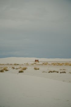 Lonely cabin surrounded by vast desert sands under an overcast sky, evoking solitude.