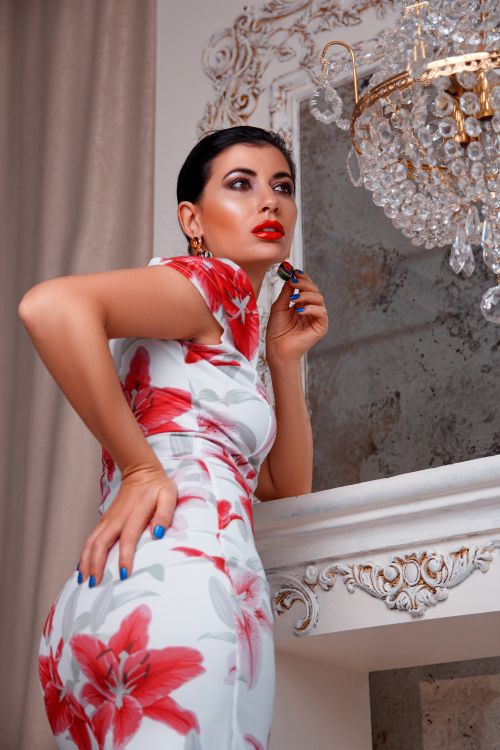 Free Stylish woman with red lips in a floral dress posing gracefully by a chandelier. Stock Photo