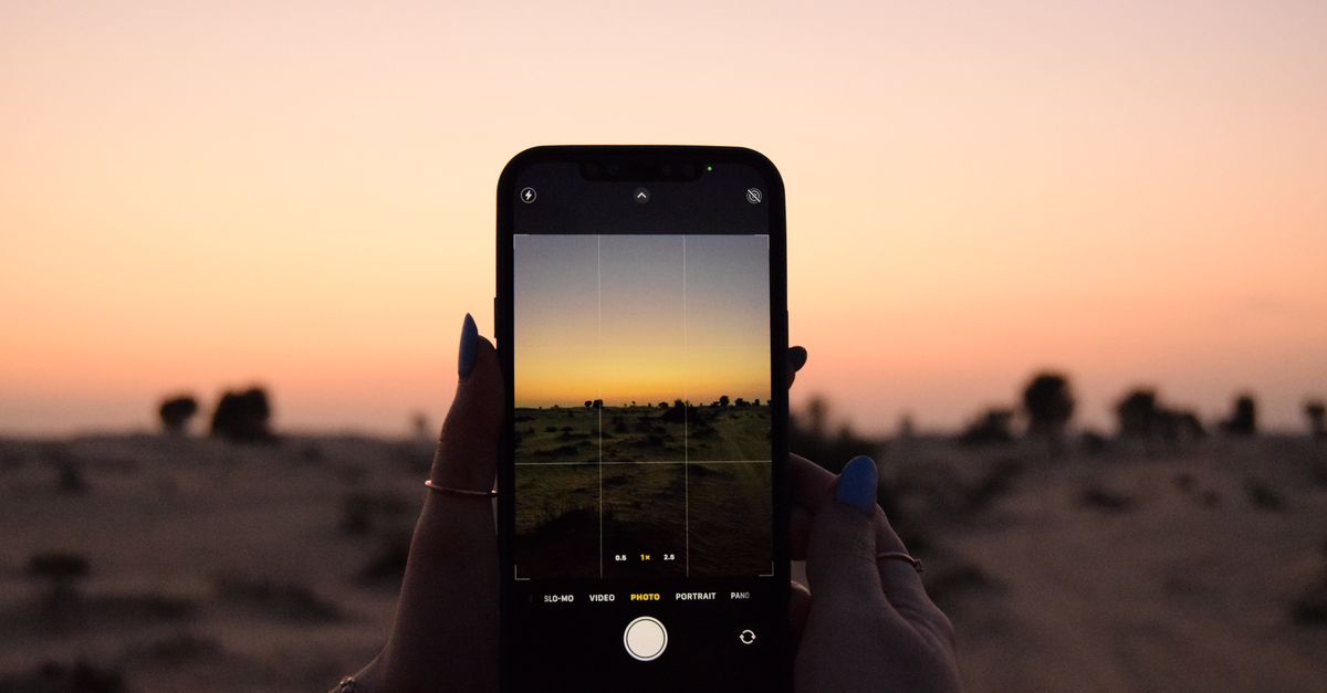 Photo by Jana Al Mubaslat Hands holding a smartphone capturing a sunset in a desert landscape.