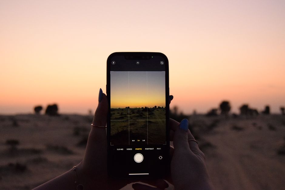 Photo by Jana Al Mubaslat Hands holding a smartphone capturing a sunset in a desert landscape.