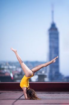 Young woman in yellow performing gymnastics on rooftop with skyscraper backdrop.