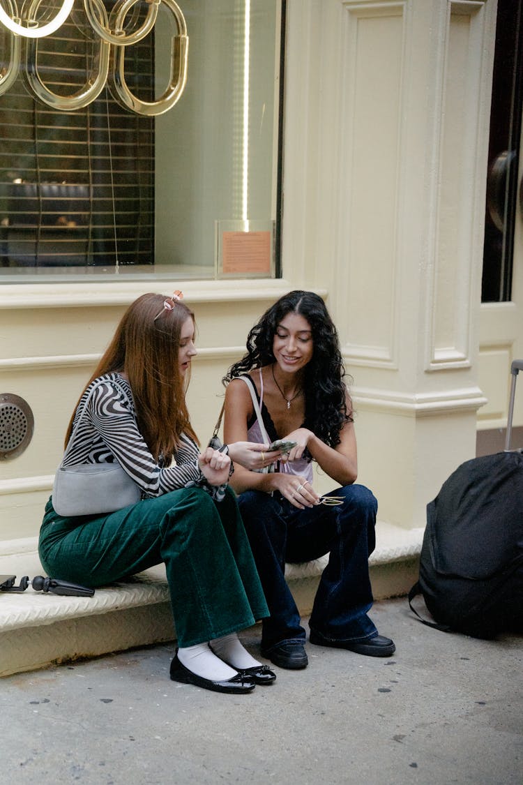 Two Young Girls Sitting On A Curb And Scrolling Through A Smartphone 