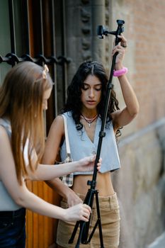 Young fashionable girls adjusting tripod equipment outdoors, embodying modern youth and creativity.