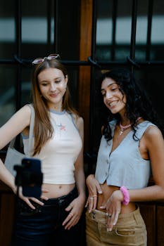 Two fashionable teenagers pose for a selfie outdoors, showcasing trendy outfits.