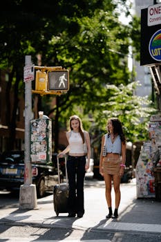 Two fashionable teenagers crossing a city street with a suitcase, enjoying a sunny day.