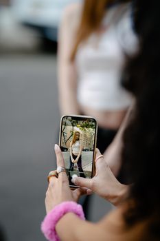 A woman capturing her friend's photo using a smartphone in a city setting, showcasing street photography.