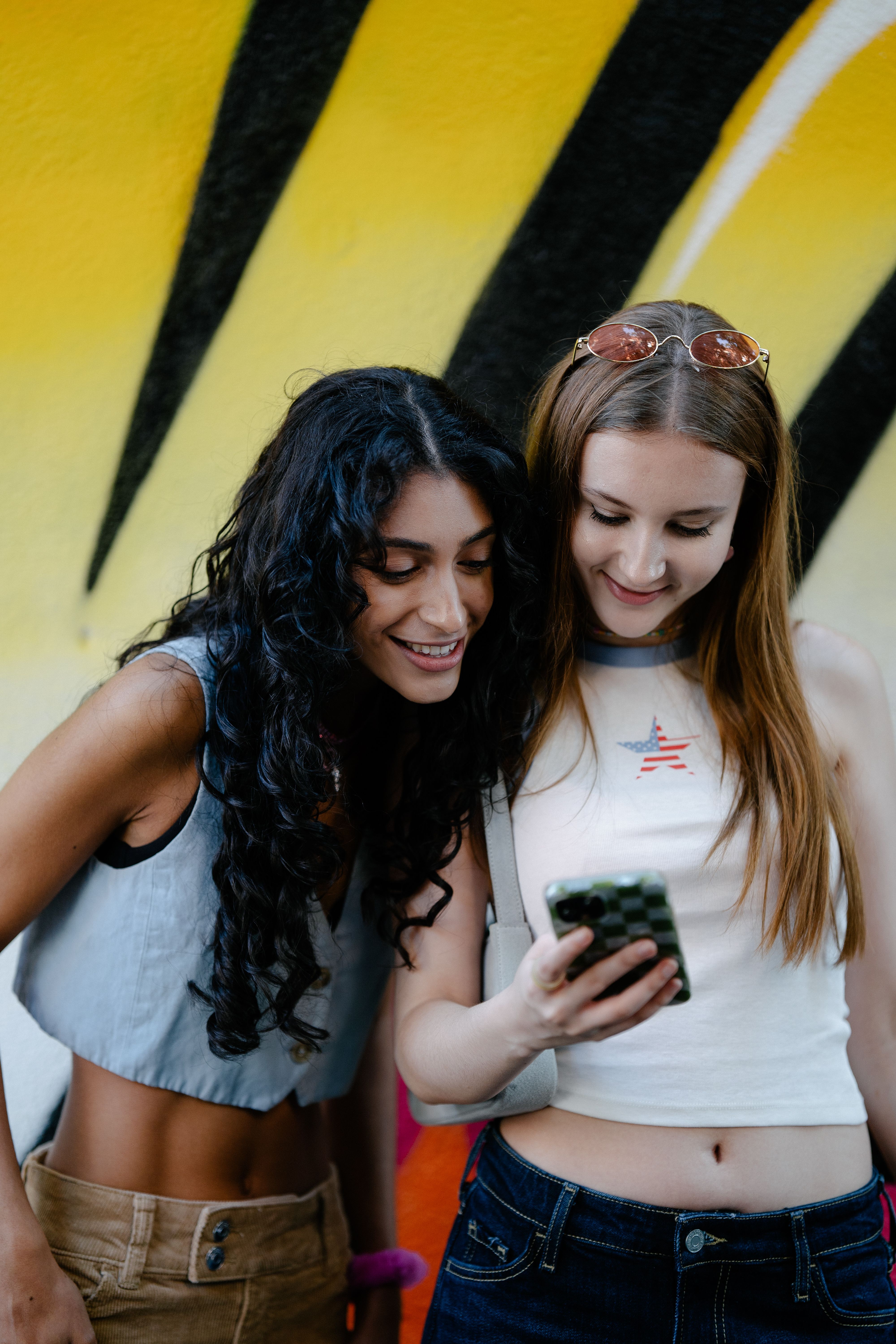Two young women smiling and looking at a smartphone against a graffiti background, symbolizing friendship and urban lifestyle.