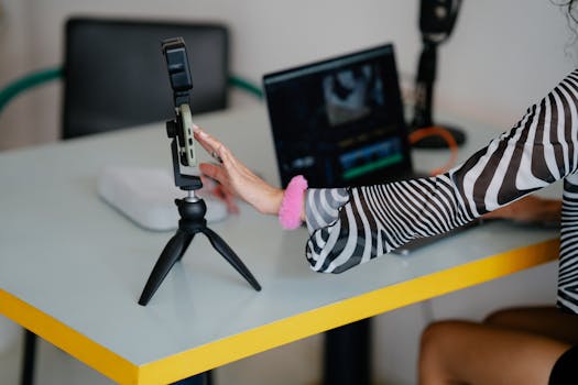 Woman adjusting smartphone on tripod with laptop setup for video editing.