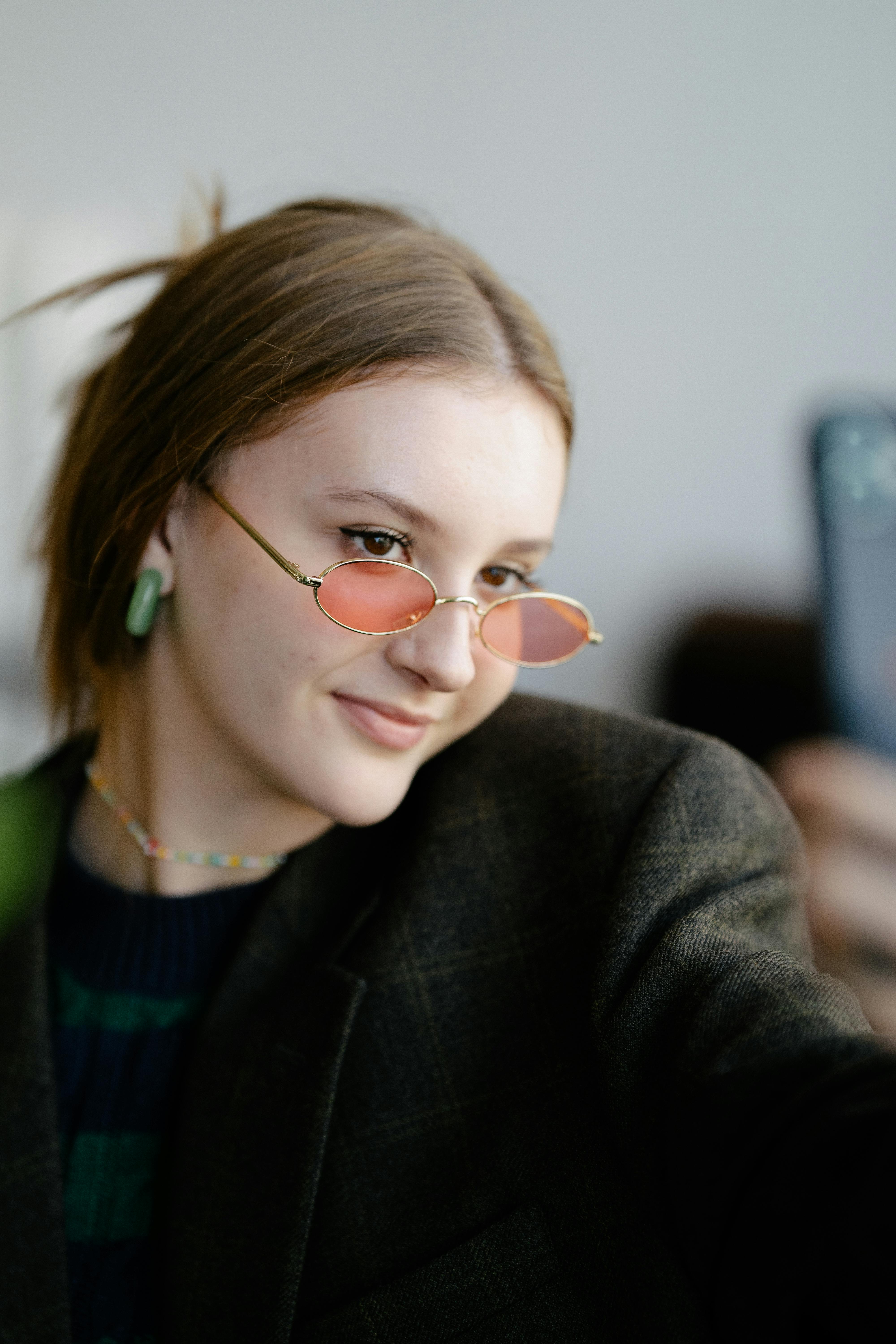 Portrait of a young woman wearing round sunglasses posing for a selfie indoors.