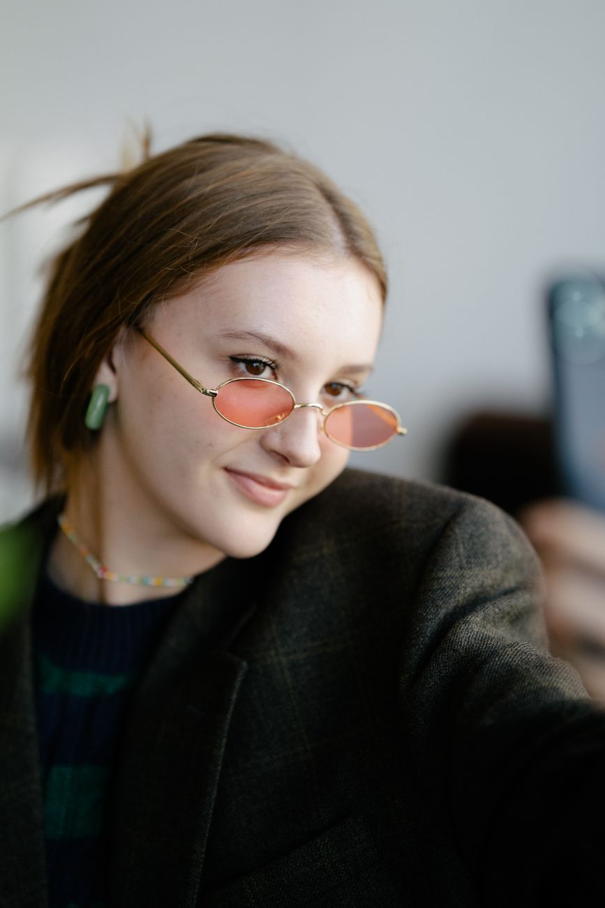 Portrait of a young woman wearing round sunglasses posing for a selfie indoors.