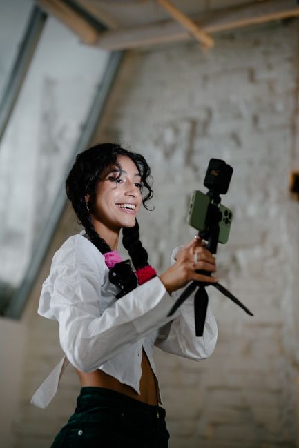 Happy woman with braids holds a smartphone mounted on a tripod indoors.