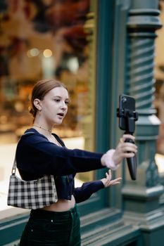 A stylish young woman takes a selfie using a smartphone stabilizer on a city street.