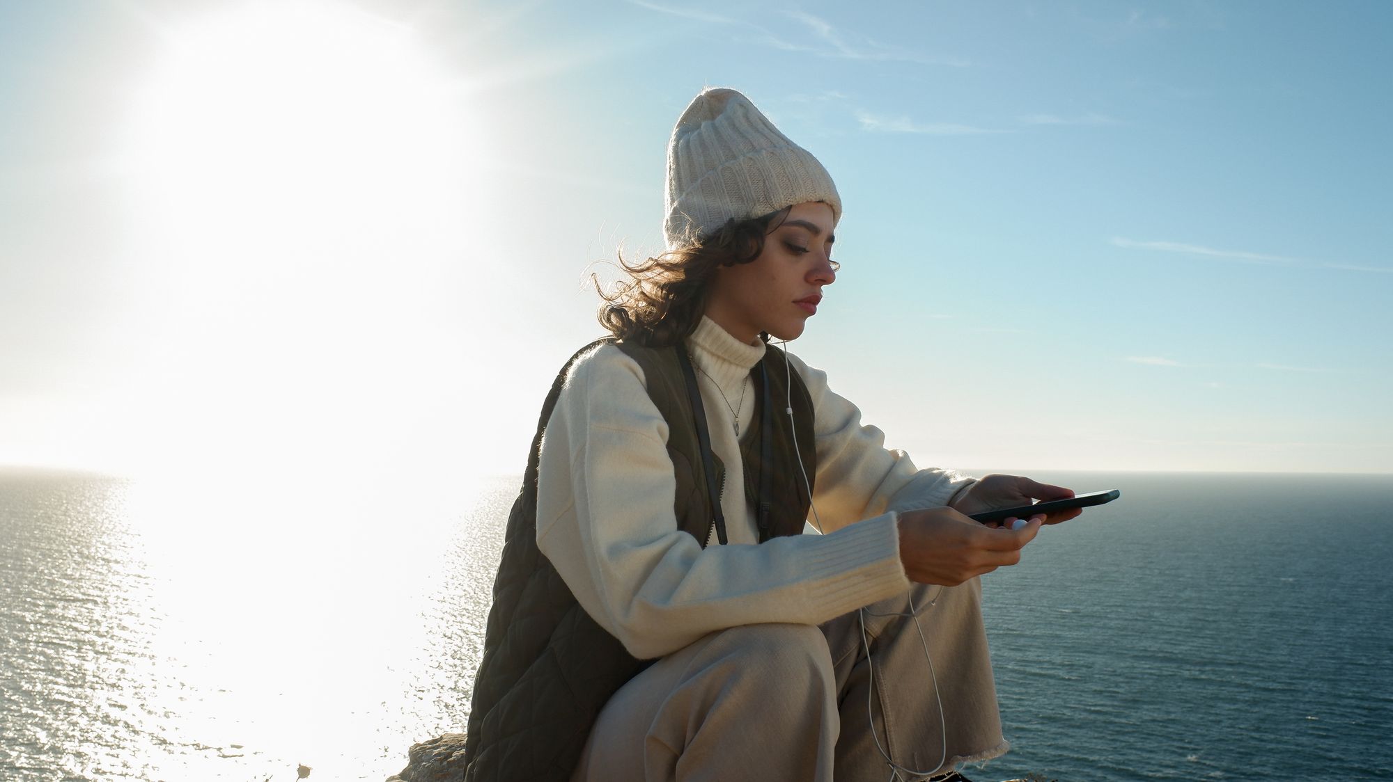 Listener with wired earphones and a phone on a coastal clifftop in low winter sun — photo by LEPTA STUDIO on Pexels