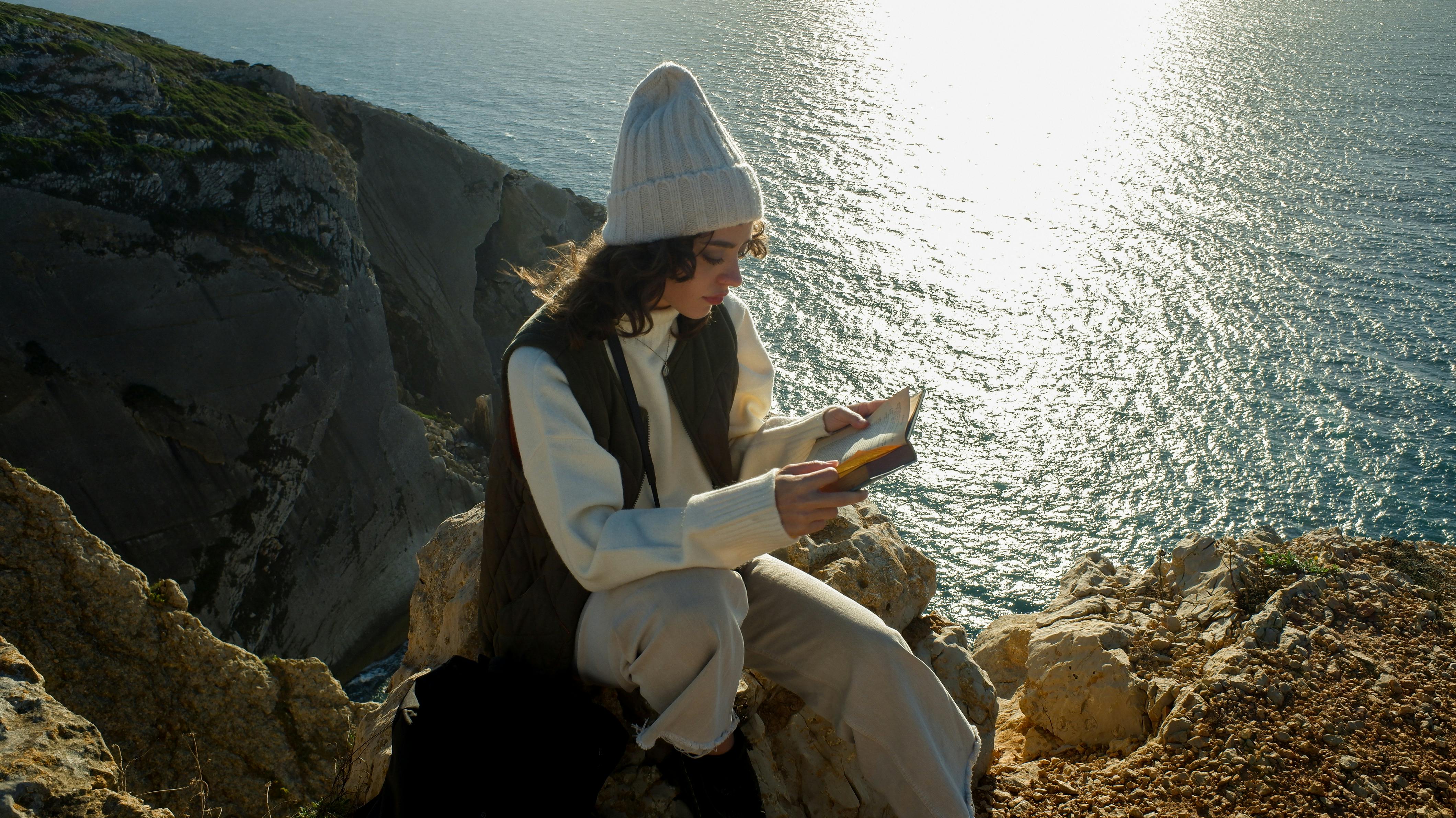 A woman sits on a Sesimbra cliff, reading a book by the ocean in Portugal.