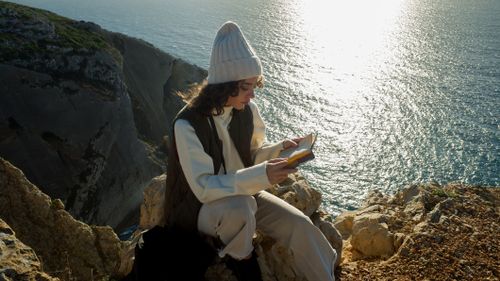 Free A woman sits on a Sesimbra cliff, reading a book by the ocean in Portugal. Stock Photo