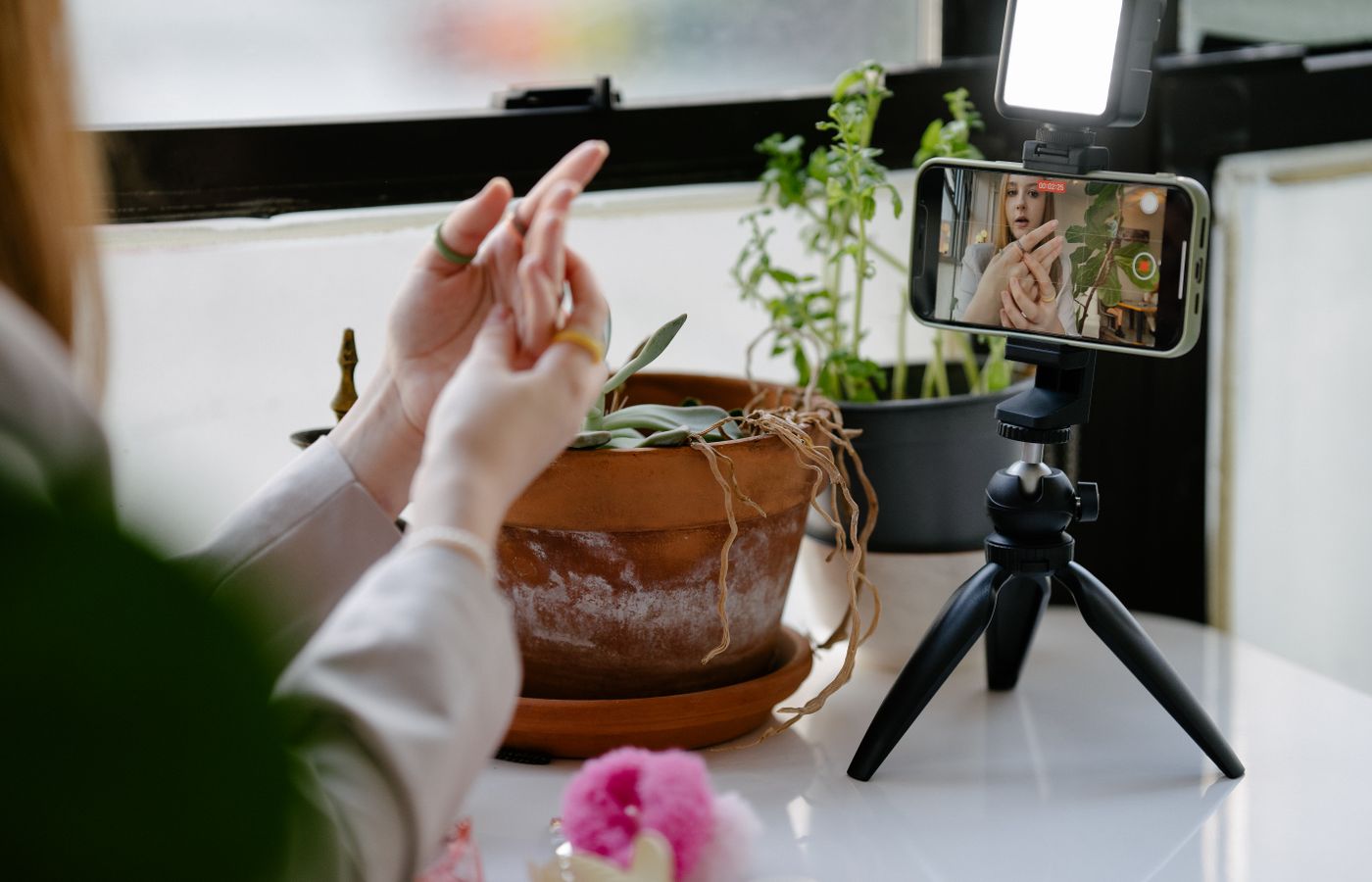 A woman uses a tripod-mounted smartphone to record a video indoors with plants in the background.