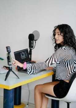 A woman recording herself with a smartphone and laptop at a desk indoors.