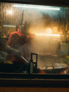 Moody silhouette of a cook seen through a steamy window in a kitchen at night, evoking warmth and mystery.
