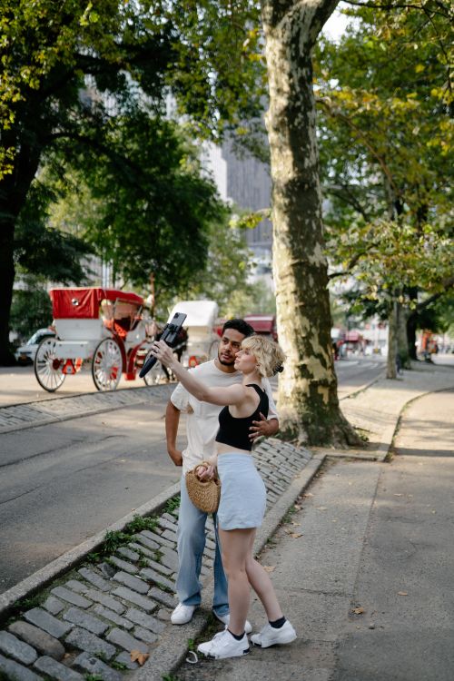 Free A couple takes a selfie in a park with a red horse carriage in the background. Stock Photo