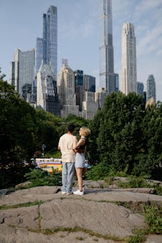 A couple stands on rocks, gazing at the New York City skyline from a park.