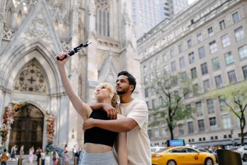Free A couple embracing and taking a selfie outside a historic cathedral in a bustling city. Stock Photo