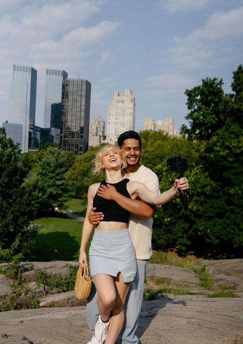 Free A joyful couple embracing and taking a selfie in a city park with skyscrapers in the background. Stock Photo