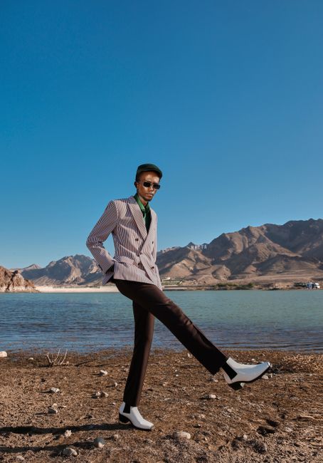Fashionable man in a suit posing outdoors by a lake with mountains in the background.