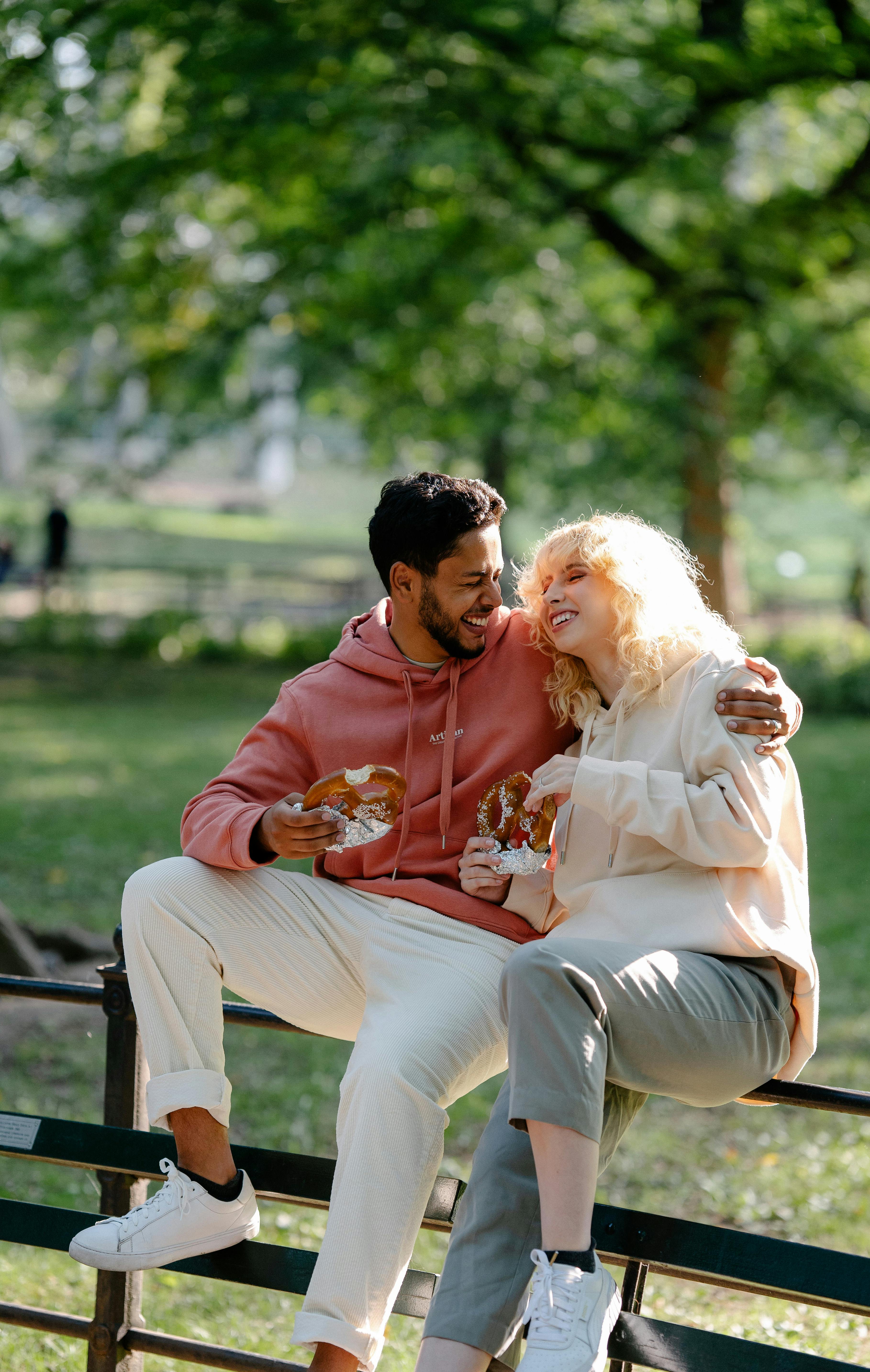 A joyful couple sitting on a park bench, sharing pretzels and laughter in a lush green setting.