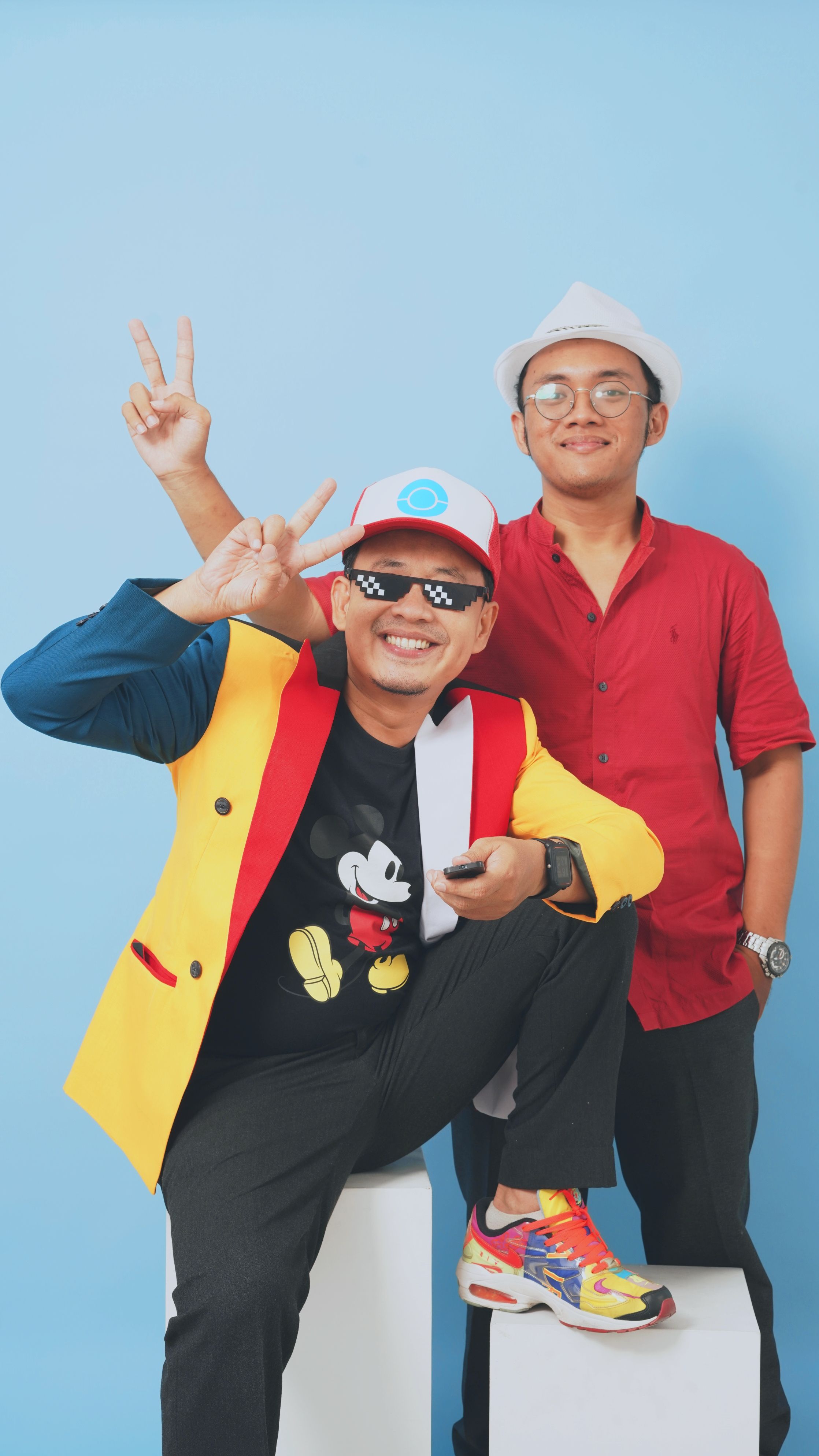 Two men in a colorful studio, posing with peace signs, fedora hats, and vibrant outfits.