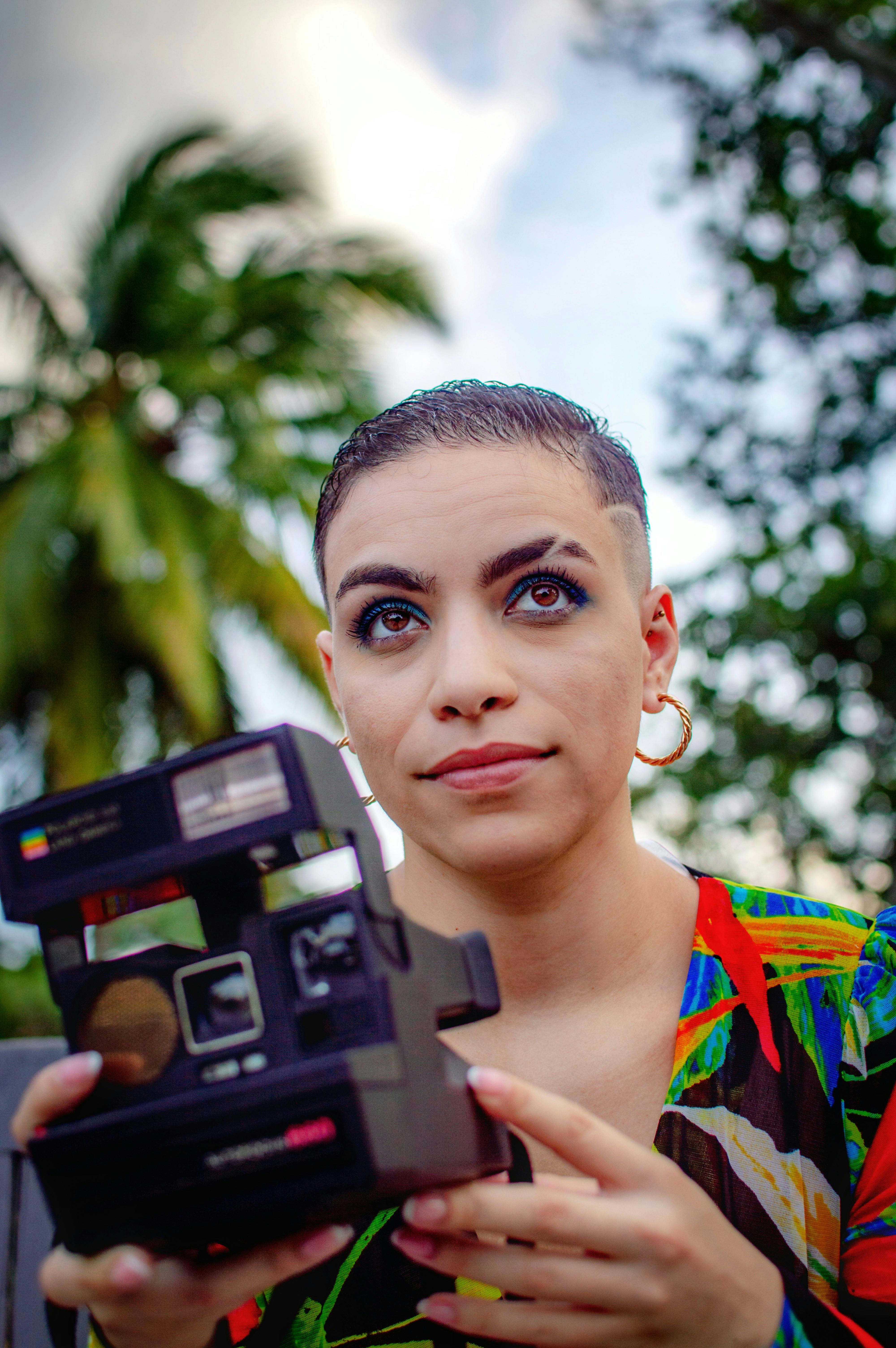 Person with short hair holding an instant camera outdoors with palm trees.